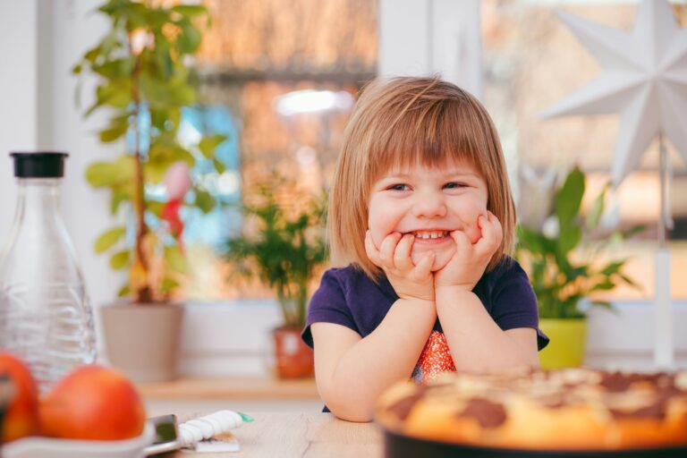 Our Blog A happy young girl smiling indoors surrounded by plants and a pastry, with natural light streaming in.