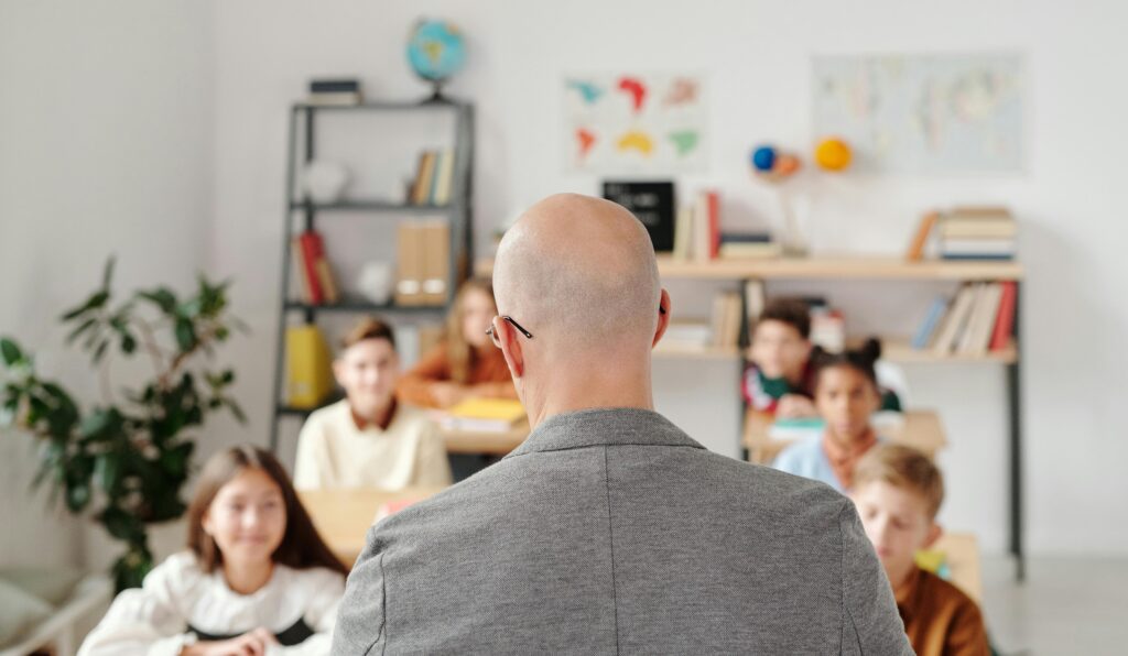 Bald teacher in a classroom engaging with attentive students sitting at desks.
