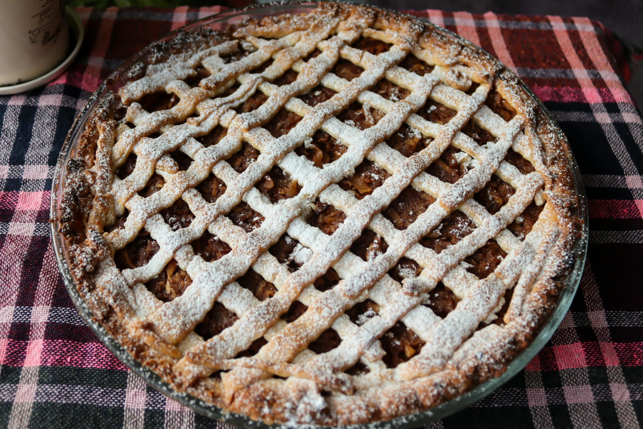 Delicious homemade apple pie with lattice crust dusted with powdered sugar on plaid tablecloth.